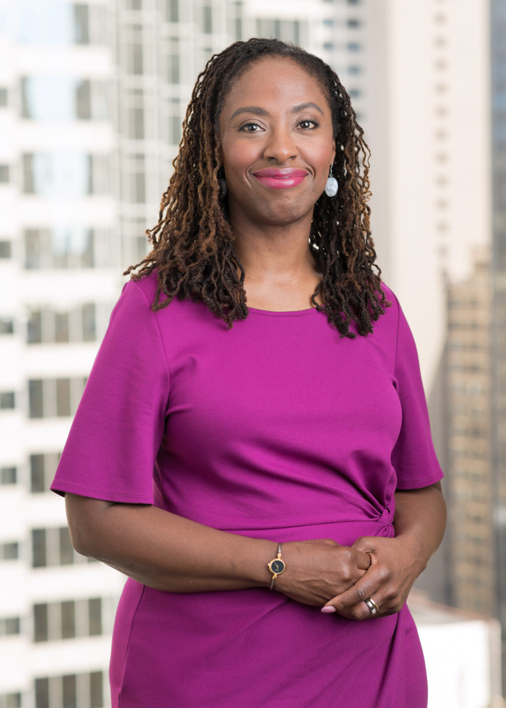 a dark-skinned woman with brown, medium-length hair in small curly locs, wearing a magenta dress gathered at the waist, hands clasped at her hip, smiling in front of a window overlooking a tall office building