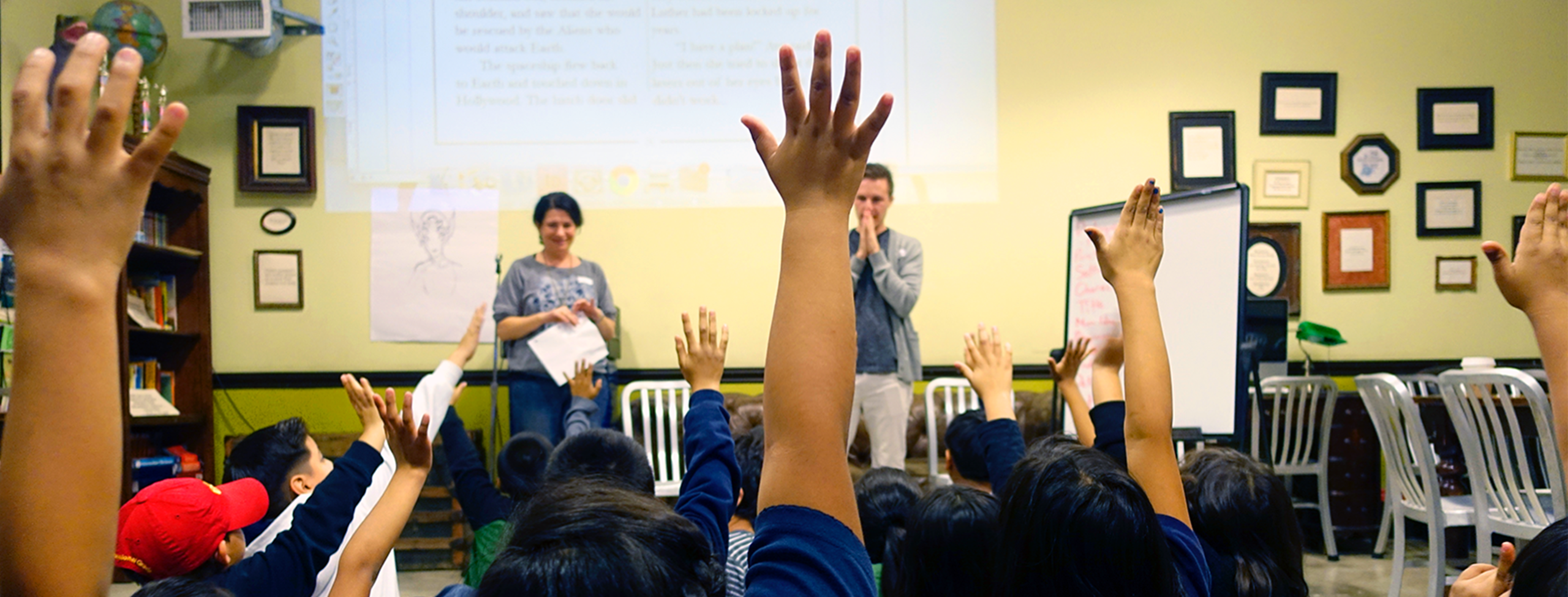 a group of about 20 children, all with black hair and medium skin, sit on the floor; the camera is behind them so we can't see their faces. about half of them are raising their hands energetically in the air. in the background, two light-skinned adults (a woman and a man) smile and seem to be deciding who to call on. they are all in a room full of silver chairs, bookshelves and numerous framed papers on the wall. a projector displays some writing on the wall behind the adults.