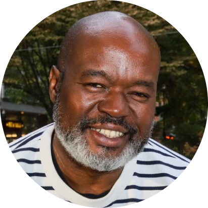 headshot of a dark-skinned man with a bald head and gray/black beard, smiling, wearing a white shirt with thin black stripes, and in front of some trees