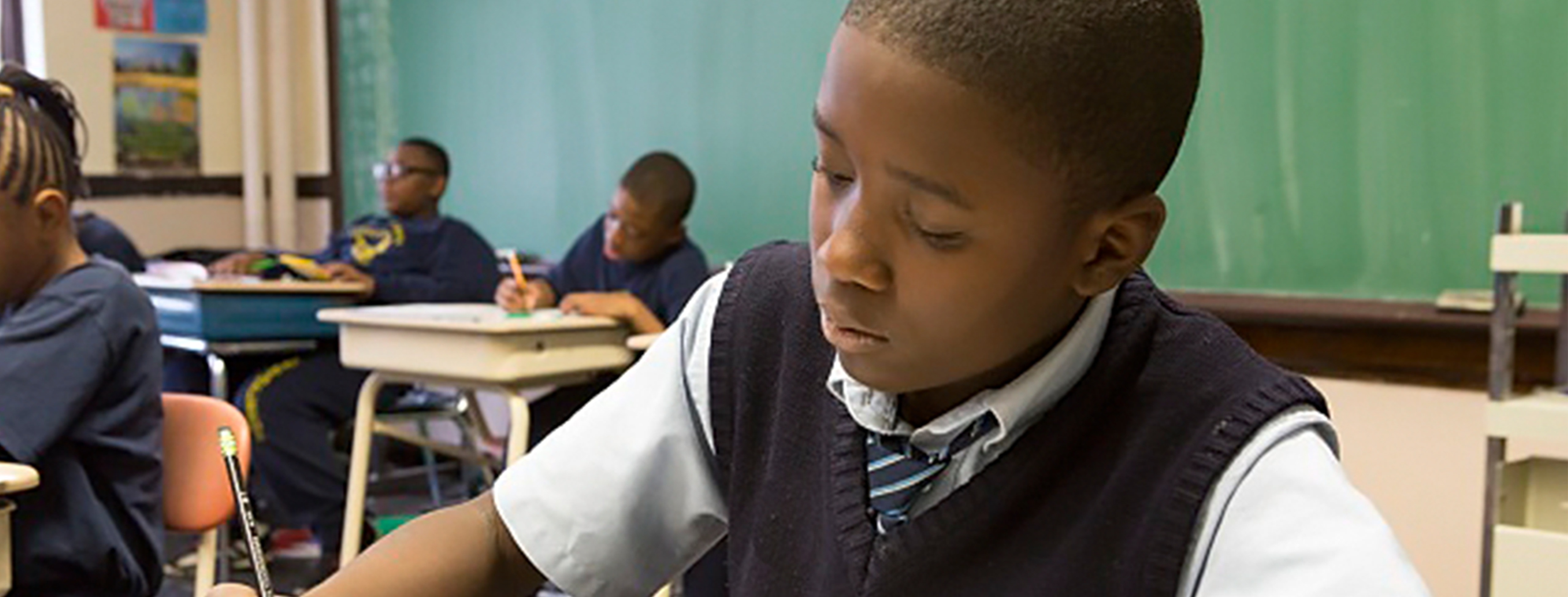 a dark-skinned boy with very short hair and a navy blue vest over a white short-sleeved shirt and tie sits at a desk in a classroom, writing with a pencil on paper. there are four other young people and a green chalkboard in the background.