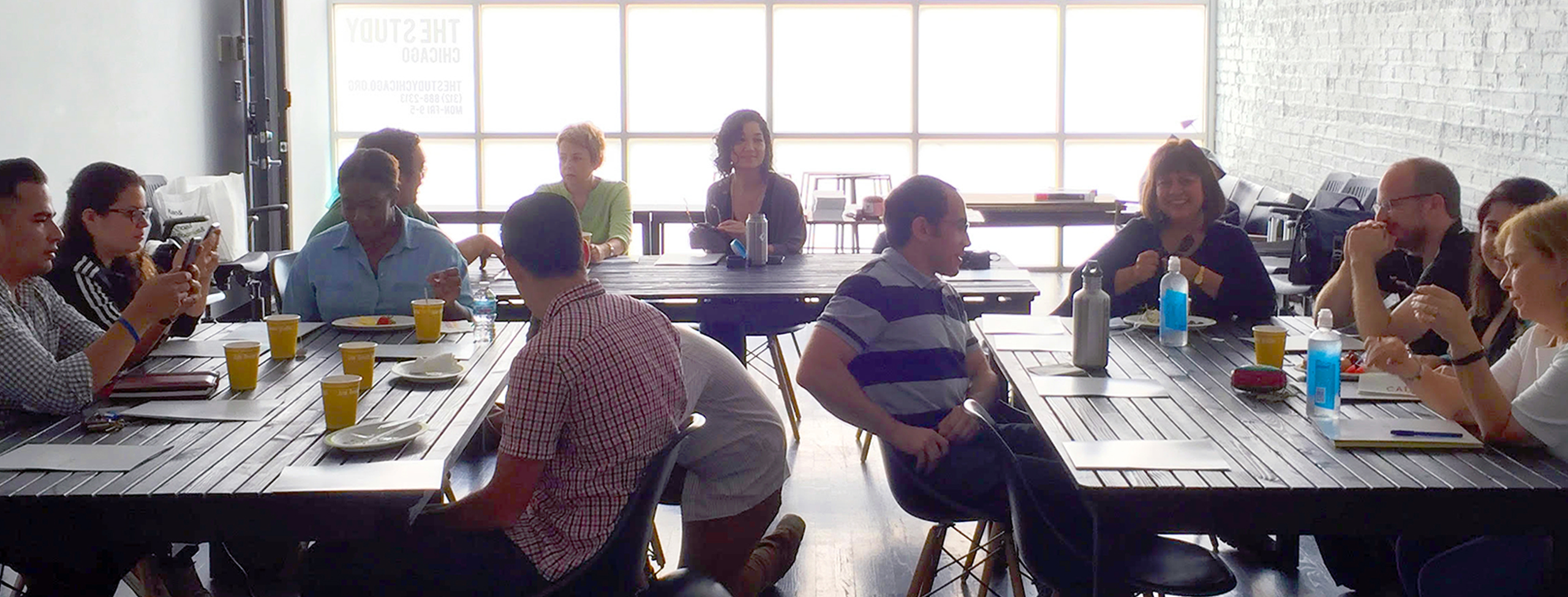 a group of 12 adults sit around three rectangular tables, each covered with papers, cups and water bottles. they are in a bright room lit by a wall of windows in the background. some are smiling or interacting, others are looking at their phones.