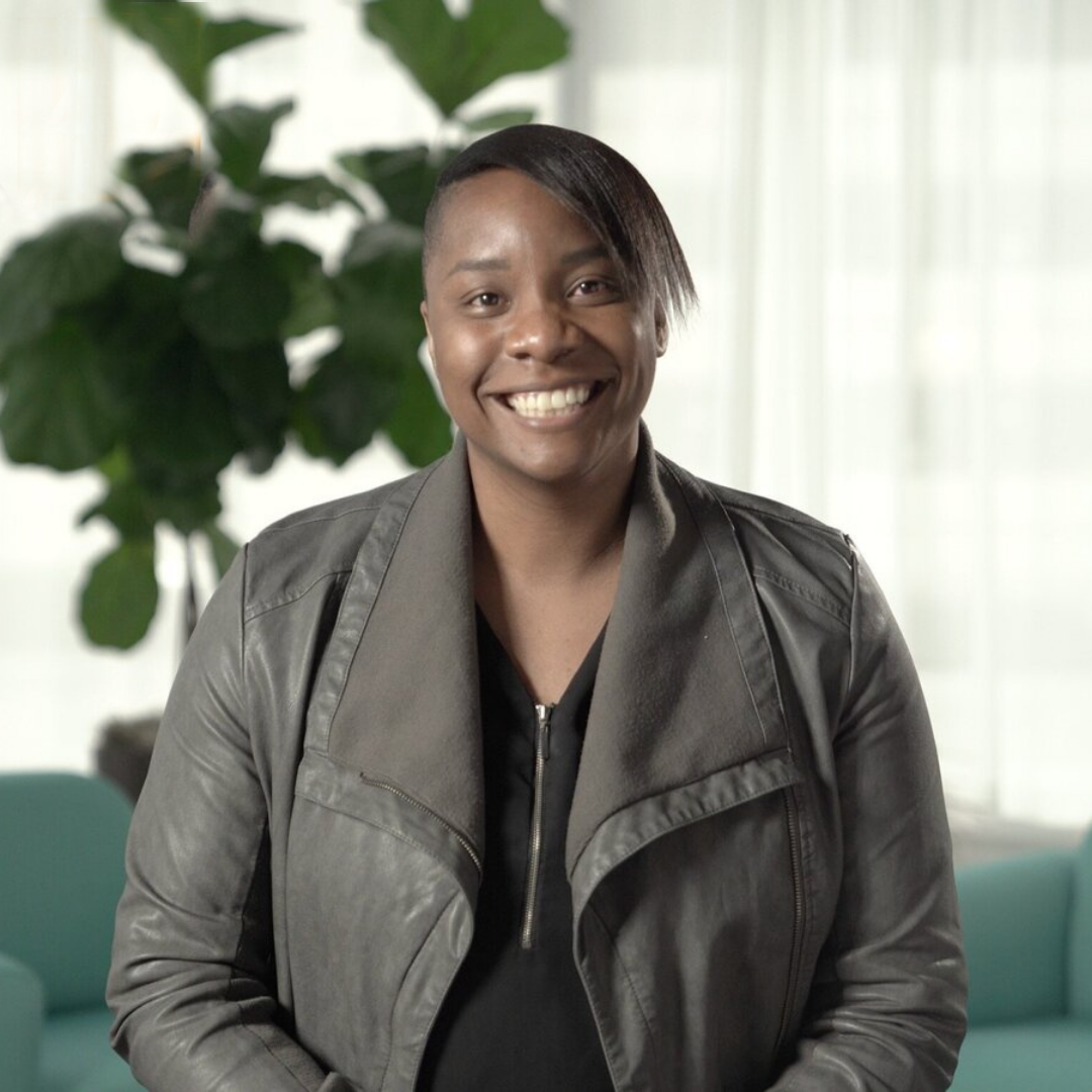 a medium-crop of a dark skinned woman with short, side swept hair and a gray leather jacket, smiling in front of a couch and plant.