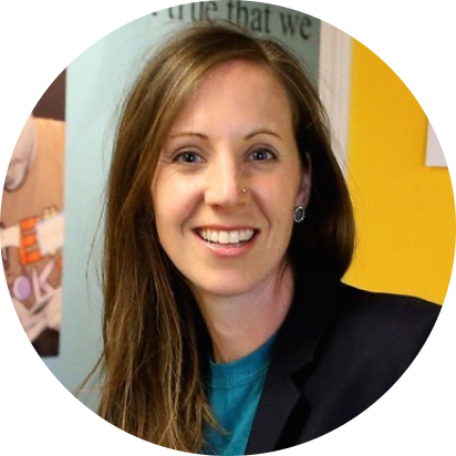 headshot of a light-skinned woman with long, straight brown hair, smiling, wearing a turquoise t-shirt and black blazer over, in front of a bright yellow and light blue wall.