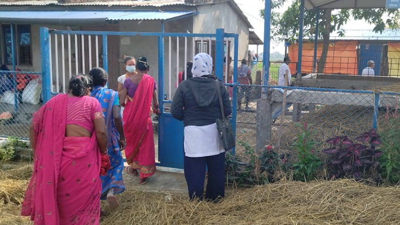 A line of three women in brightly-colored saris face away from the camera; their heads are down as they walk through a gate and into a yard. A person in a face mask appears to greet them at the threshold. In the backyard are two low buildings and in the foreground is a pile of hay.