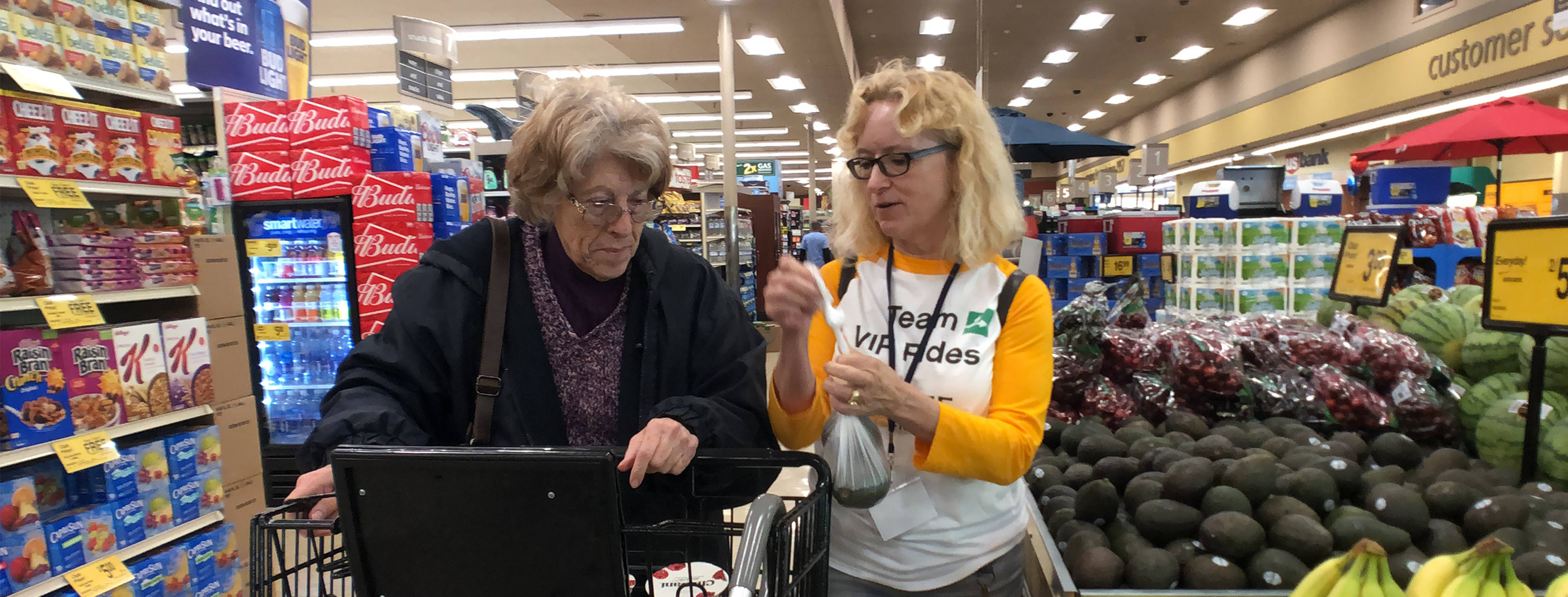 two women face the camera in the middle of a grocery store. both are light-skinned; the one on the left has gray hair and glasses and pushes a shopping cart. the one on the right has blond hair and a bright white t-shirt with yellow baseball-style sleeves. she is holding a plastic bag with produce in it.