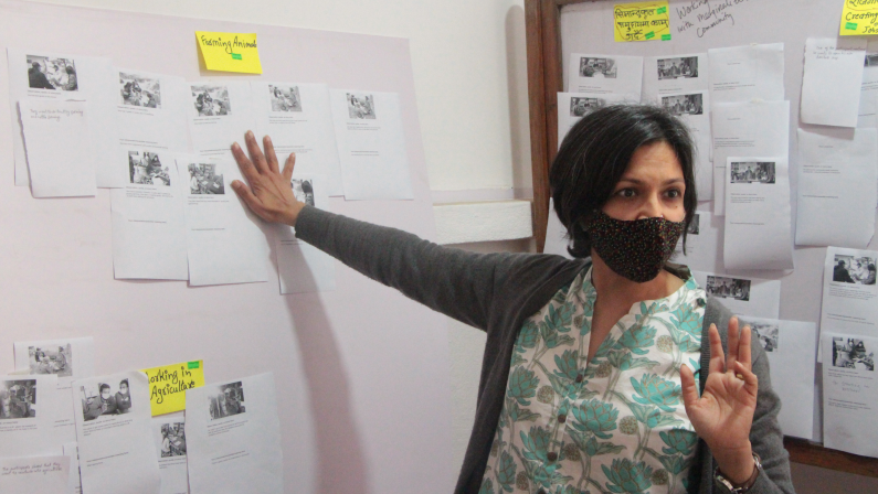 A woman stands surrounded by white walls that have small sheets of white paper as well as yellow post-it's stuck to them. She wears a mask over her nose and mouth. One hand rests on the wall behind her and the other is raised in a gesture as she looks intently off-camera to the right. 