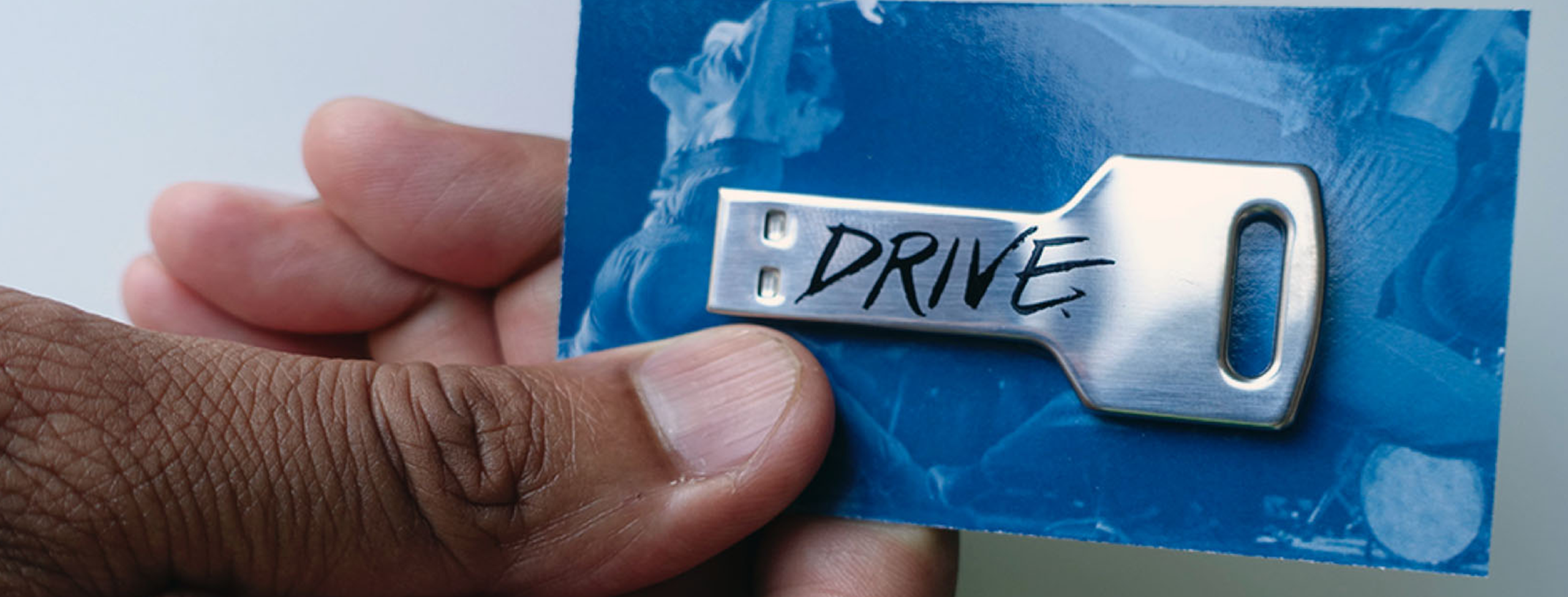 a medium-skinned hand holds a blue card which has a silver key-shaped USB drive attached to it. the USB says the word "DRIVE" in aggressive font. the blue card is a duotone photo of dancers in evocative poses.