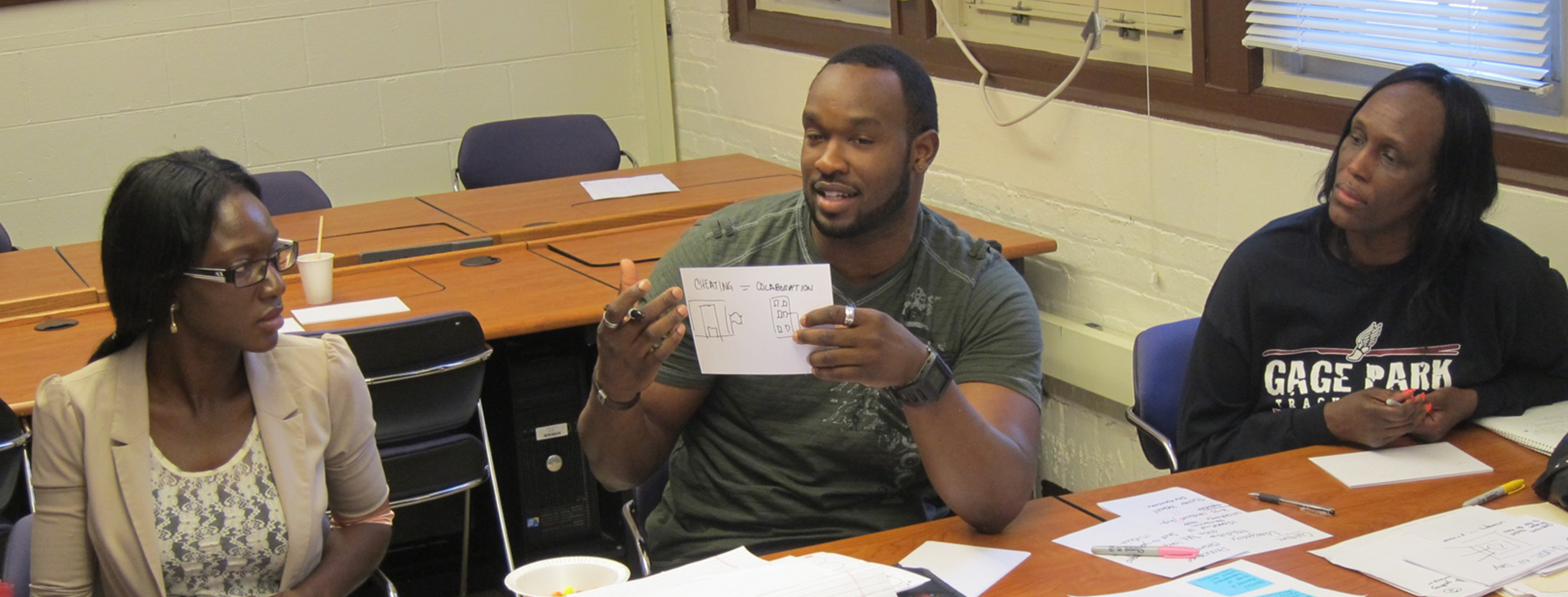 three dark-skinned adults sit at a long table that is covered with papers. in the center is a man holding up a half sheet of paper, upon which there is a drawing done with Sharpie. the women on his left and on his right look at his drawing with interest.