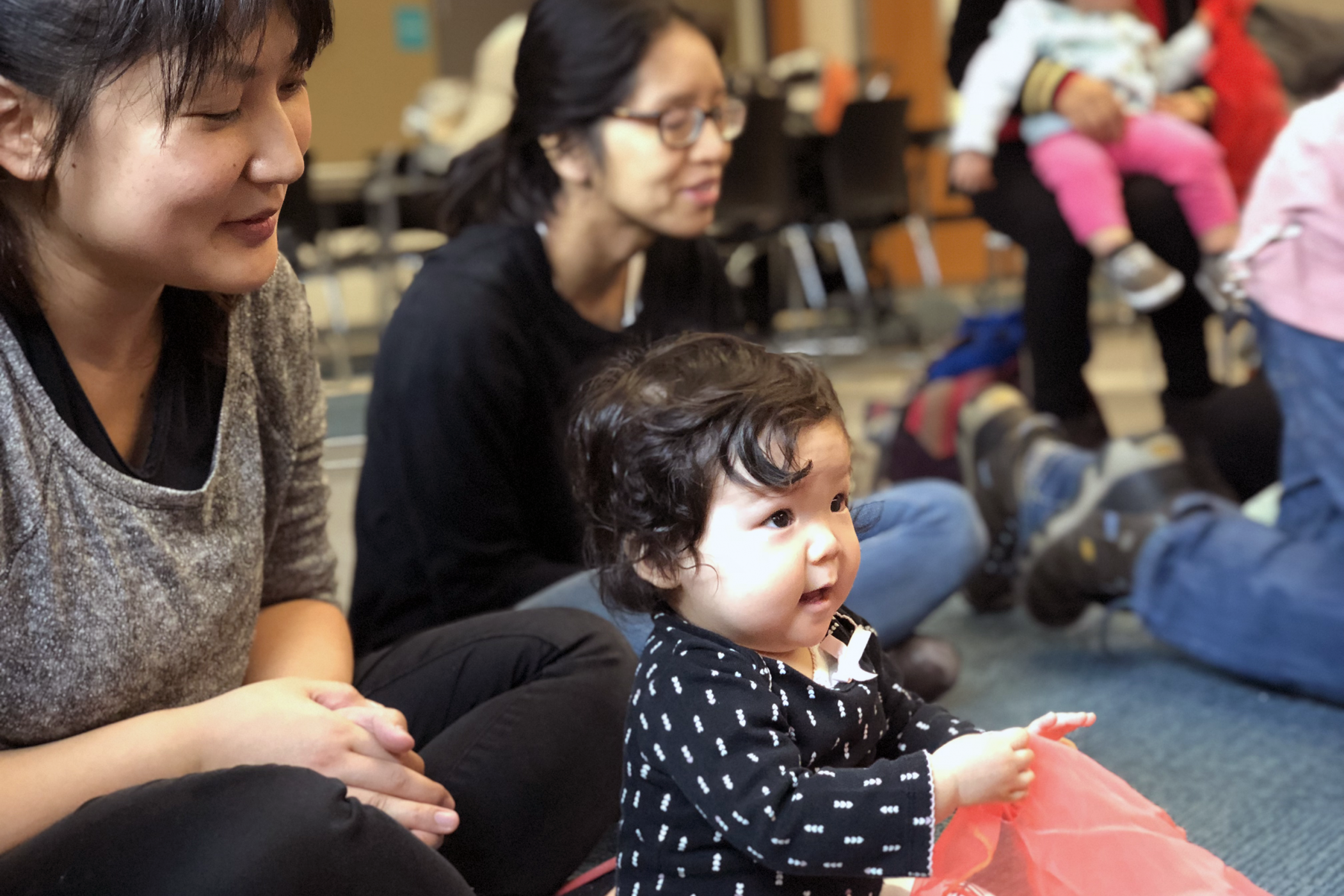 An Asian baby sits on the floor smiling with caregivers and other adults around her