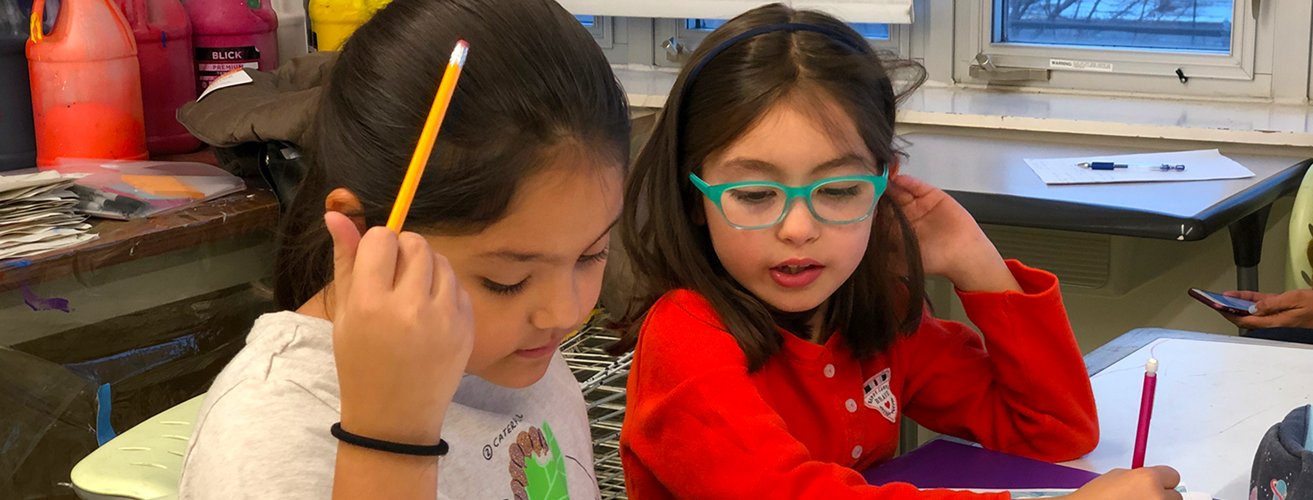 two elementary school-aged girls sitting next to each other at a table, looking down at one's work. the one on the left holds a yellow pencil and wears a gray t-shirt; her hair is in a ponytail. on the right the girl wears a bright red shirt and turquoise-framed glasses. her hair is straight and brown, down with a thin black headband.