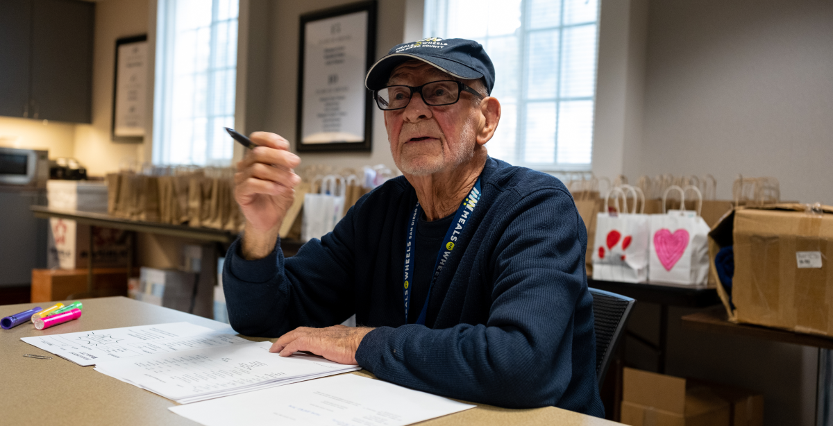 Photo of an older man looking up and holding a pen, while seated at a table with papers in front of him.