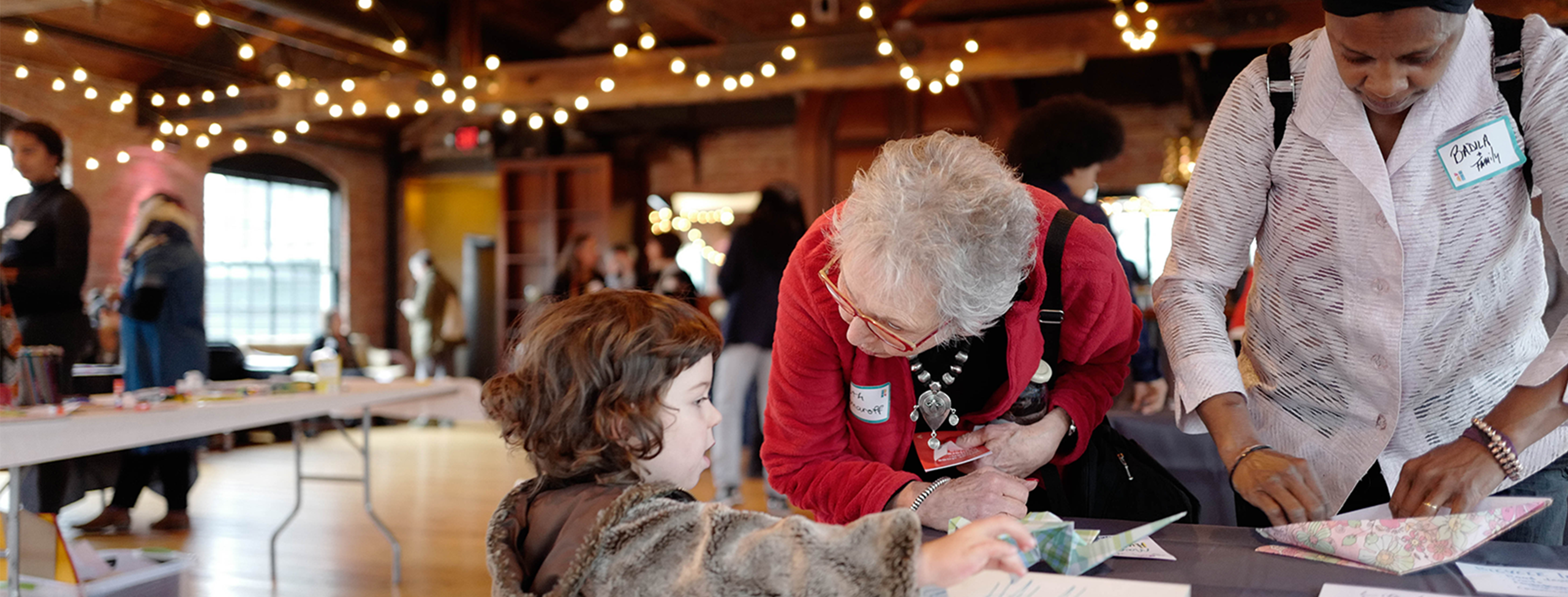 a woman with gray hair and light skin in a red sweater leans forward over a table, her head turned towards a young light-skinned girl in a furry brown coat. next to them, a medium-skinned woman in a white shirt and black head wrap folds a piece of paper. they are in a room with tall ceilings, exposed wooden rafters and string lights.