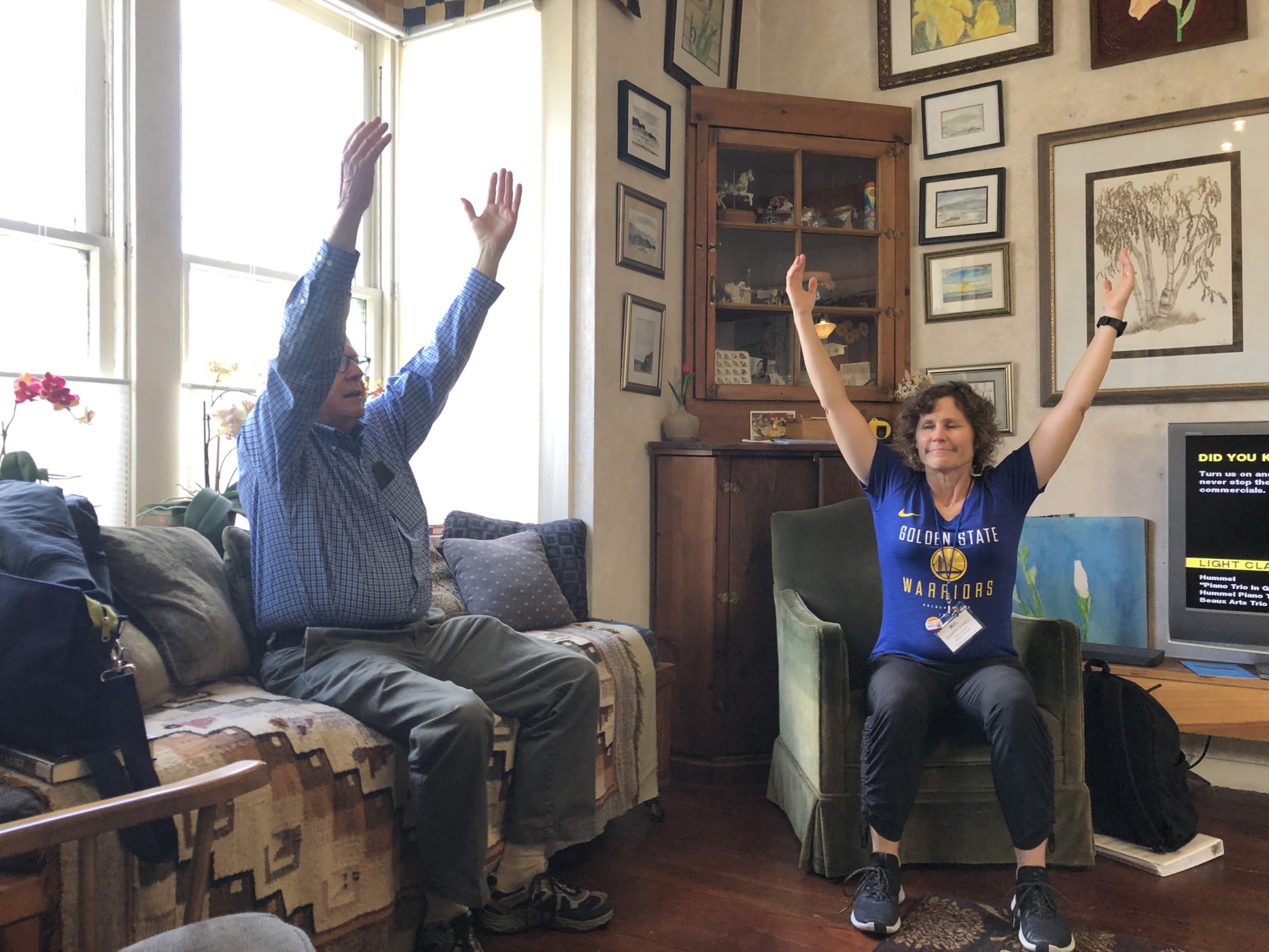 Two older adults sitting on couches in a living room, with their arms raised straight up in the air